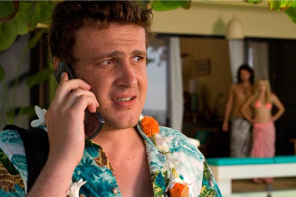 Jason Segel em uma cena no Havaí, falando ao telefone com uma expressão angustiada, com um casal ao fundo