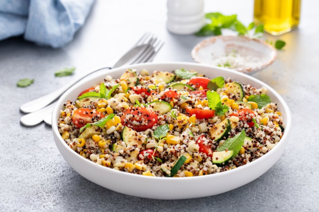 Recipiente branco com quinoa cozida, tomates-cereja, milho-verde, feijão-fradinho e folhas de hortelã em cima de uma mesa cinza