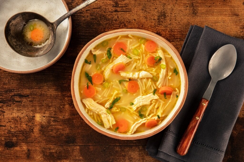 Sopa de frango com macarrão, cenoura e legumes em recipiente claro ao lado de colher em pano preto e concha em recipiente claro