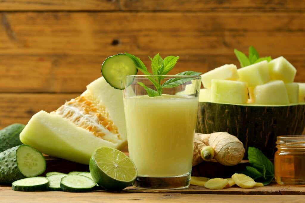 Suco de melão com pepino, gengibre, limão e hortelã servido em copo, cercado pelos ingredientes frescos sobre uma mesa rústica de madeira