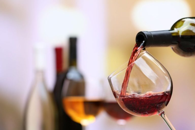 Vinho tinto derramando em taça de vidro com raças e garrafas desfocadas ao fundo