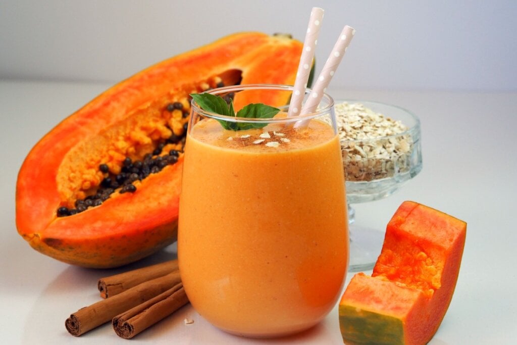 Vitamina de mamão com aveia e canela servido em copo de vidro, decorado com hortelã e canudos, ao lado de mamão, pau de canela e aveia em flocos