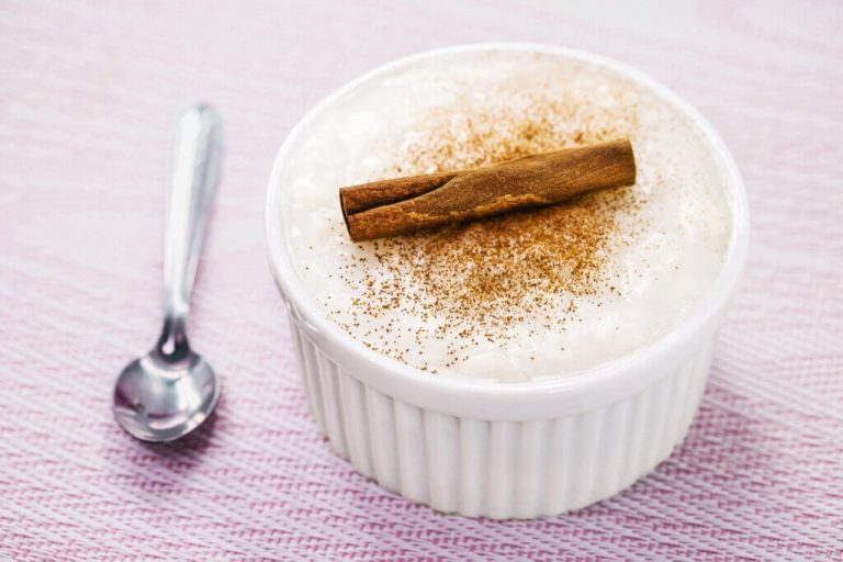 Ramequin com arroz-doce polvilhado com canela em pó e decorado com canela em pau sobre jogo americano rosa e com uma colher ao lado