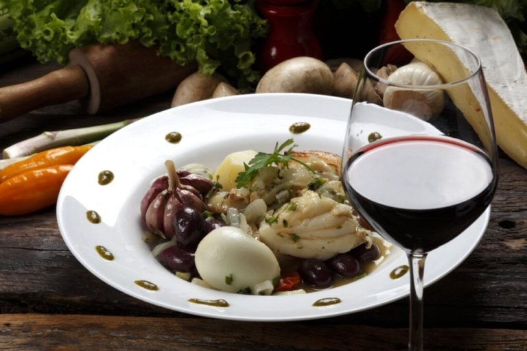 Prato com bacalhau acompanhado de legumes e taça de vinho tinto em uma mesa rústica com ingredientes ao fundo