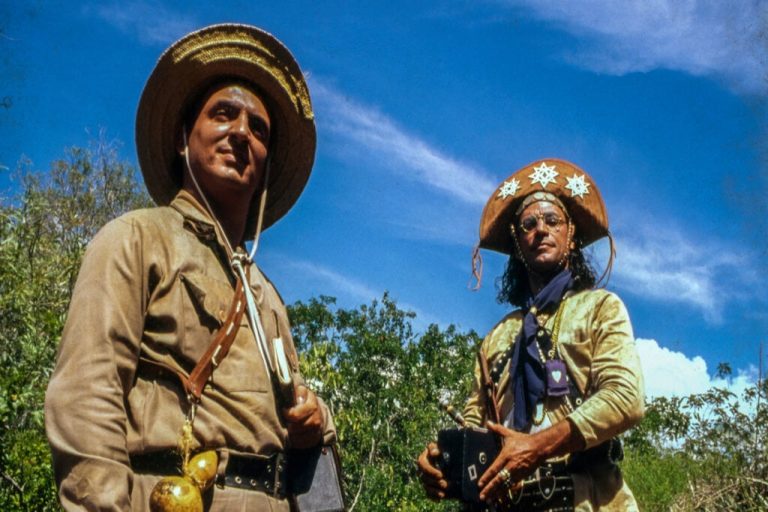 Dois homens vestidos com roupas de cangaceiro no meio da mata. Um deles está com uma câmera antiga na mão e o outro com um caderno. 