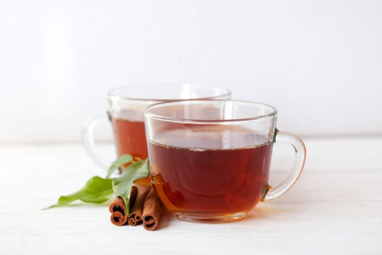 duas canecas de vidro com chá marrom. canelas em pau e folhas de chá verde ao lado