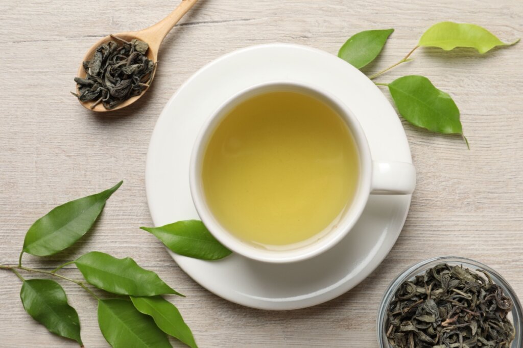 Chá verde servido em xícara branca e com folhas secas e frescas de chá verde ao redor em cima de mesa de madeira