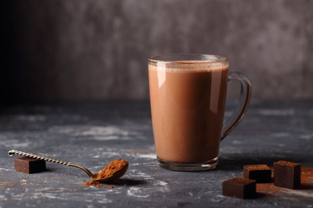 caneca de vidro com chocolate quente e colher ao lado com cacau e cubos de chocolate