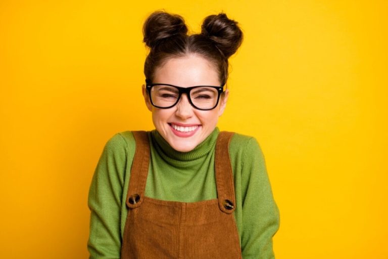 Mulher com dois coques na cabeça e usando óculos de grau, sorrindo em frente a um fundo amarelo