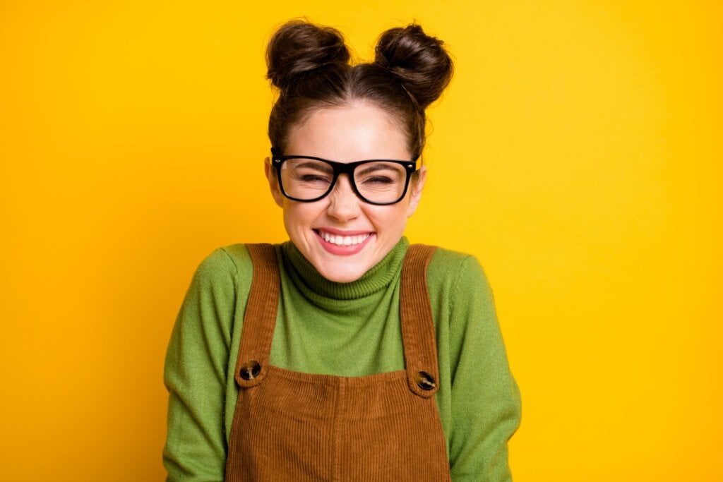 Mulher com dois coques na cabeça e usando óculos de grau, sorrindo em frente a um fundo amarelo