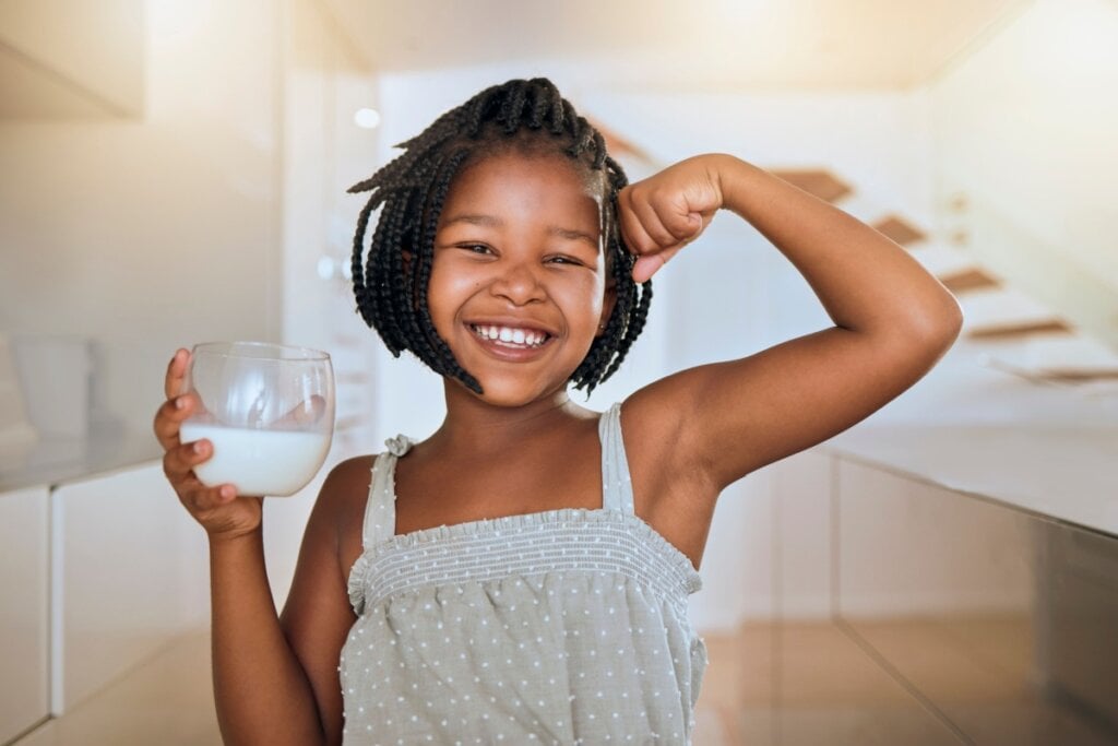 Menina com o cabelo curto trançado, usando uma regata cinza e com um copo de leite na mão