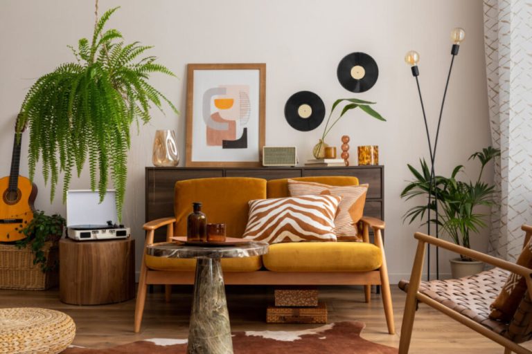 Sala com cadeiras em madeira e estofado amarelo-escuro com almofadas com estampa listrada branco e laranja, com plantas ao redor e tapete com estampa de animal e atrás das cadeiras um móvel em madeira escura com quadros e objetos de decoração