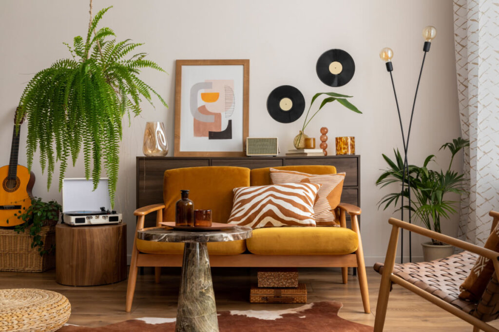 Sala com cadeiras em madeira e estofado amarelo-escuro com almofadas com estampa listrada branco e laranja, com plantas ao redor e tapete com estampa de animal e atrás das cadeiras um móvel em madeira escura com quadros e objetos de decoração