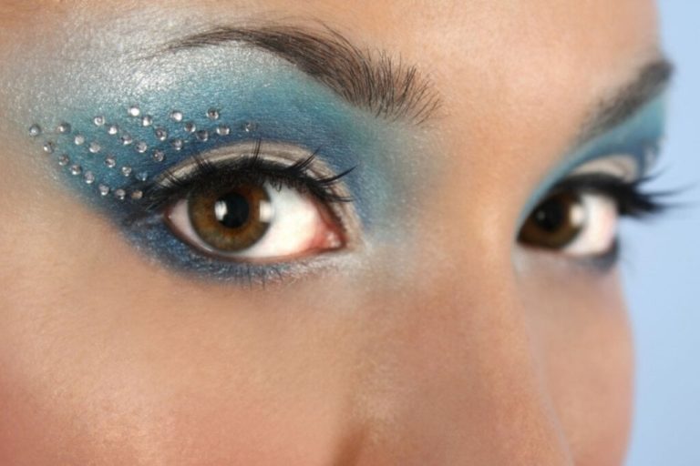 Close-up de olhos com sombra azul cintilante em tons oceânicos, cílios destacados e pedrinhas brilhantes aplicadas ao redor