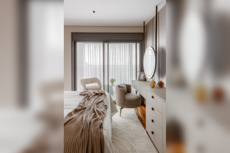 Quarto com cama coberta por manta de tricô bege, tapete claro e poltrona próxima à escrivaninha com espelho redondo iluminado