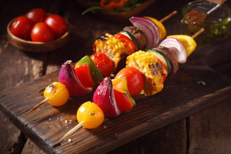 Espetinho de milho com, tomate, pimentão, cogumelo, tomate e cebola servido em tábua de madeira em cima de mesa de madeira