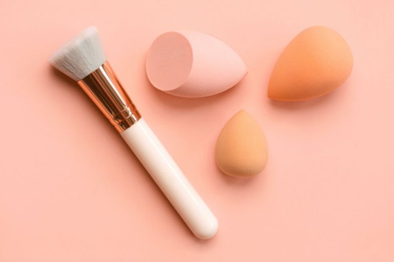 Pincel branco ao lado de três esponjas de maquiagem e sobre um fundo rosa-claro