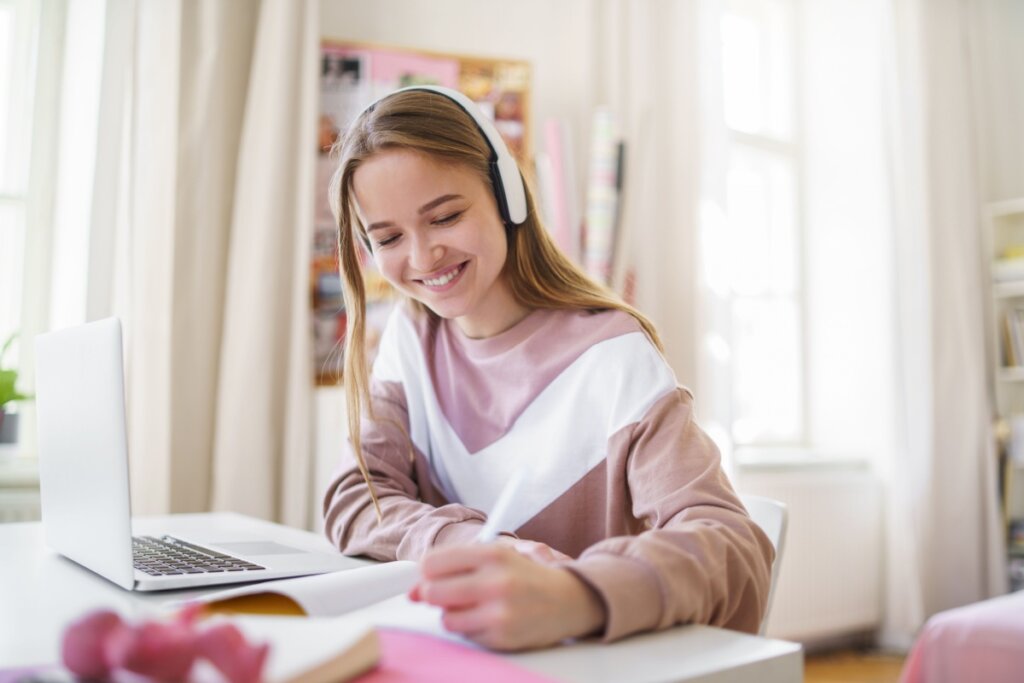 menina loira sorrindo, com fone de ouvido e escrevendo em caderno. notebook aberto à frente dela
