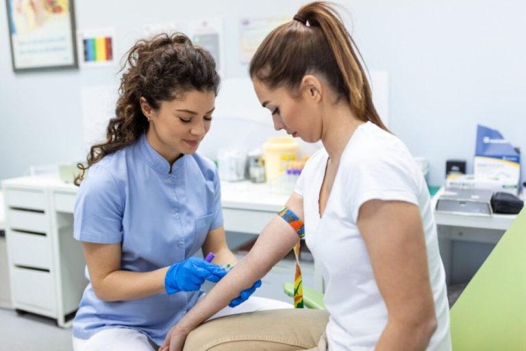 Mulher com o cabelo cacheado preso no alto da cabeça, usando jaleco de enfermeira, com outra mulher usando camiseta branca e calça bege e com o cabelo preso em rabo de cavalo em consultório médico, enfermeira está preparando a paciente para doação de sangue