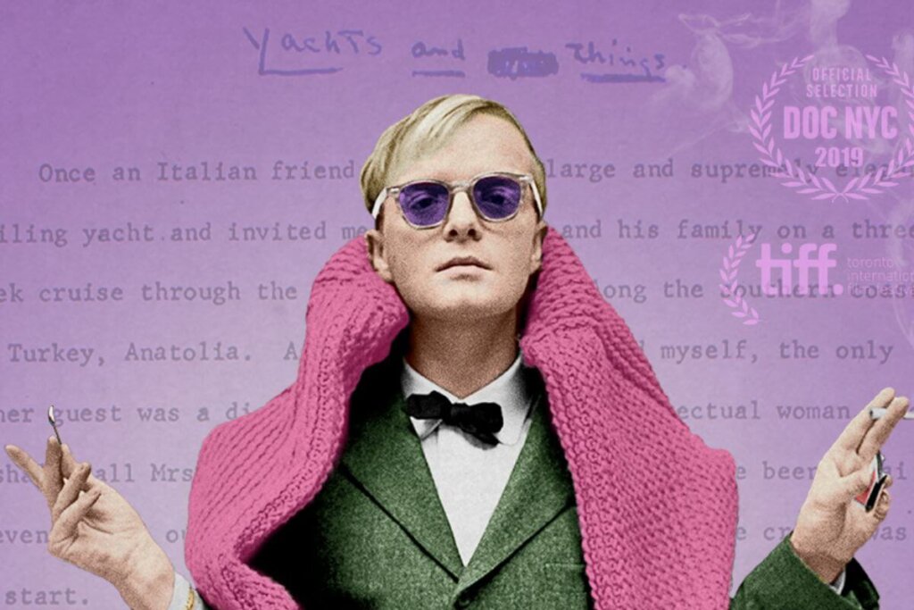 Imagem de um homem loiro, com o cabelo curto, usando um óculos escuro roxo e um terno verde, com camisa branca e gravata borboleta preta e com coberto por uma echarpe rosa em fundo roxo