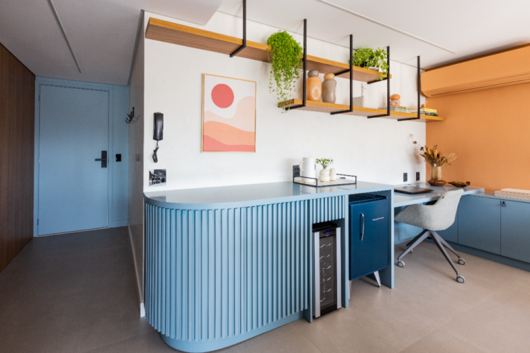 imagem de bar em casa com mesa e geladeira pequena em tons de azul, parede branca com prateleiras e planta e cadeira cinza ao fundo em home office
