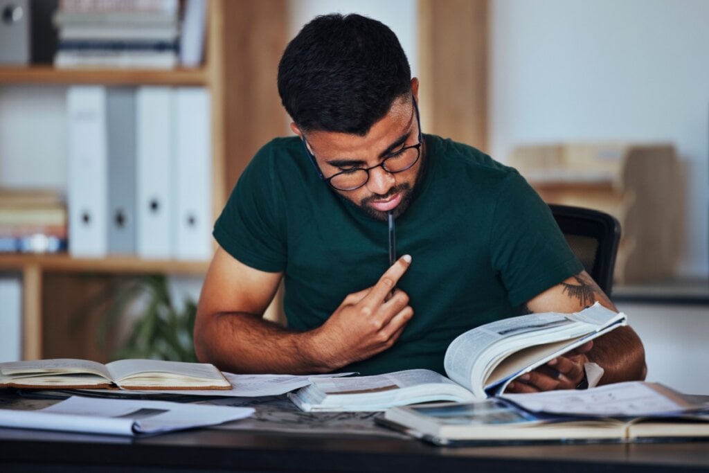Jovem com cabelo curto, usando óculos preto e camiseta verde-escura, com caneta encostada no rosto estudando diversos livros