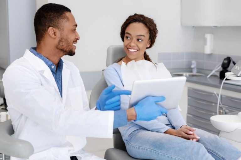 Jovem com o cabelo preso e usando roupas em tons de azul sentada na cadeira do dentista, e ao lado está um homem com jaleco de dentista e com um tablet na mão