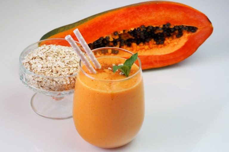 Copo de vitamina de mamão e aveia, aveia em grãos em recipiente ao lado e mamão cortado ao meio ao fundo