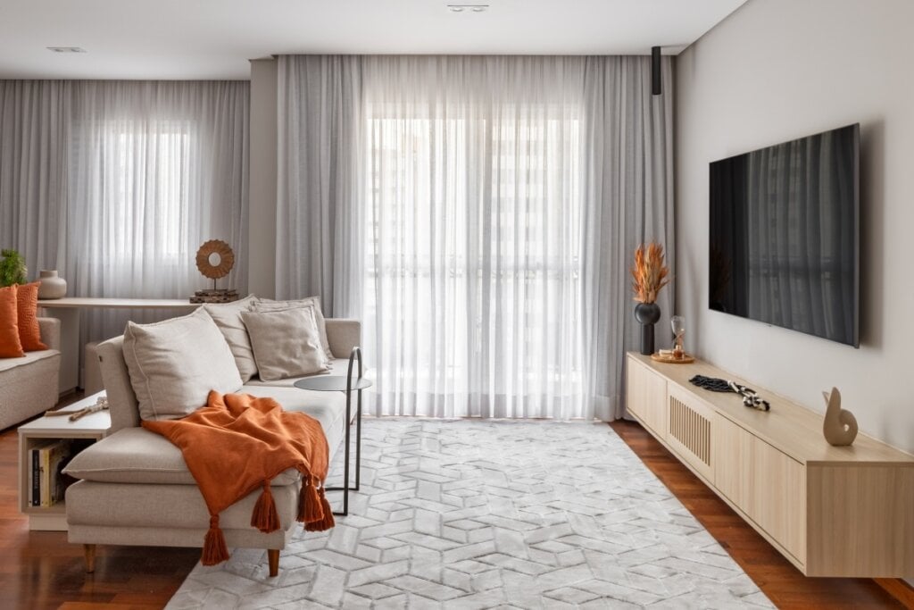 Sala de estar com sofá claro decorado com almofadas e manta laranja, cortinas translúcidas e tapete geométrico sobre piso de madeira