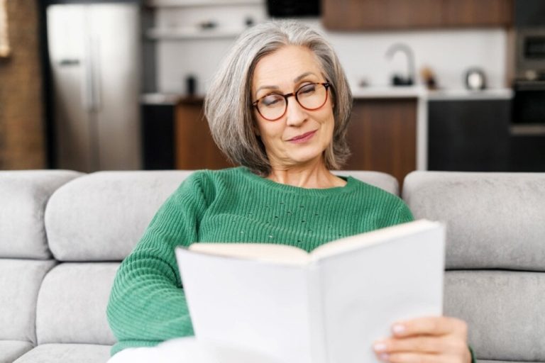Mulher com cabelo curto grisalho, usando suéter verde lendo um livro sentada no sofá