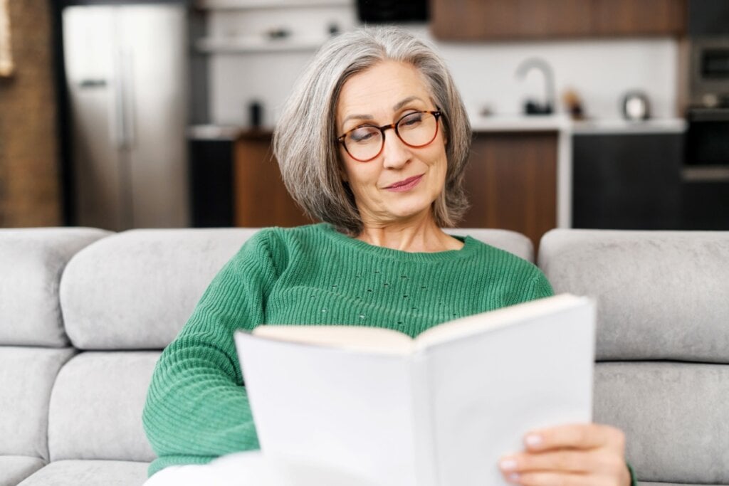 Mulher com cabelo curto grisalho, usando suéter verde lendo um livro sentada no sofá