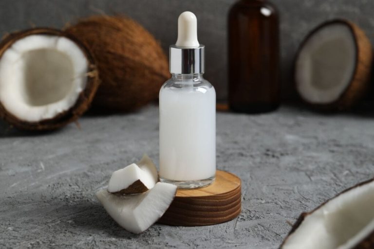 Vidro com óleo de coco em cima de um pedaço de madeira redondo e um pedaço de coco ao lado; no fundo, há um coco aberto.
