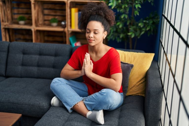 Mulher sentada em posição de meditação sobre o sofá, com olhos fechados e mãos em prece