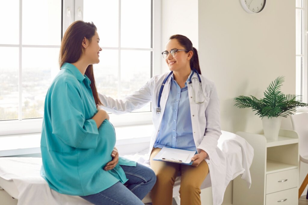 Paciente grávida com o cabelo liso, solto e usando uma camisa verde ao lado da médica que está com o cabelo preso, usando óculos e camisa azul e um jaleco branco