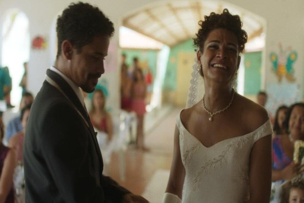 Imagem de um casal, o homem tem o cabelo curto e barba e está usando terno, e a mulher tem o cabelo cacheado e preso em véu e está usando um vestido de noiva