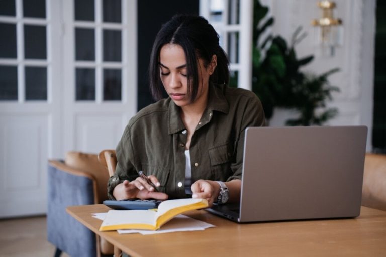 Jovem com cabelo preto e usando camisa social verde fazendo calculo na calculadora e no caderno.