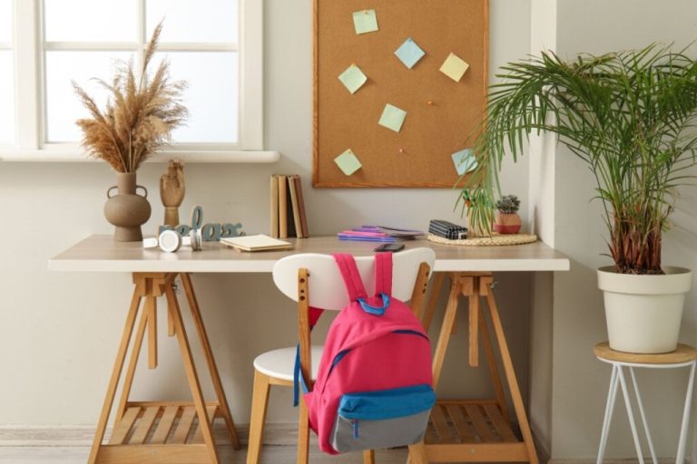 Mesa de madeira em frente a uma cadeira, com mochila pendurada e, acima dela, um quadro com post-its