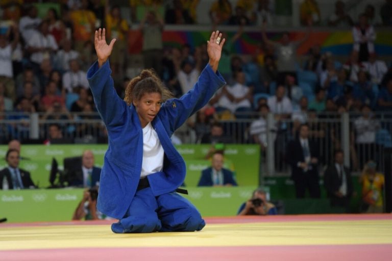 Mulher com roupa de judô azul e camiseta branca ajoelhada e com as mãos para cima em tatame de competição das Olimpíadas 