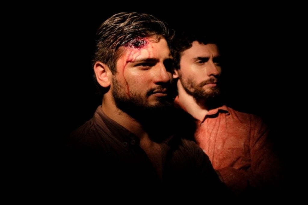 Imagem de dois homens em um local escuro, ambos com o cabelo curto e barba, um está usando uma camisa marrom e tem um ferimento na cabeça e o outro está usando uma camisa laranja