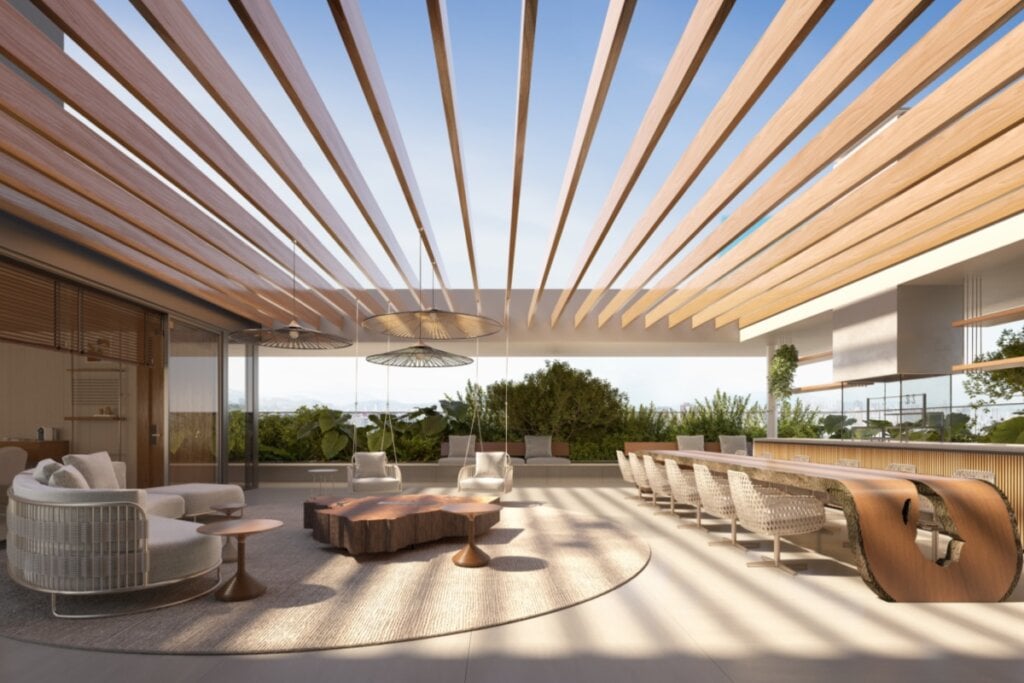 Terraço com cobertura vazada de vigas de madeira, sofás arredondados em fibra natural, mesa de centro com tampo orgânico em madeira maciça e ao fundo uma ilha gourmet