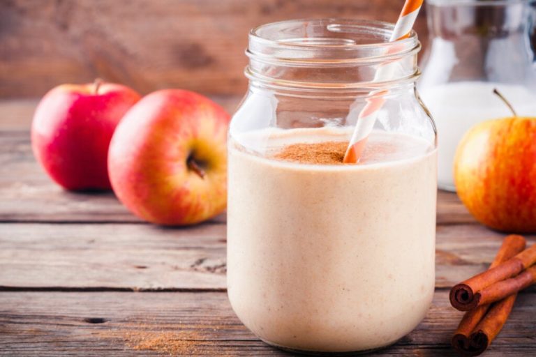 Copo com vitamina de maçã e canela em pó por cima. Ao fundo, há maçã e, ao lado do copo, canela em pau