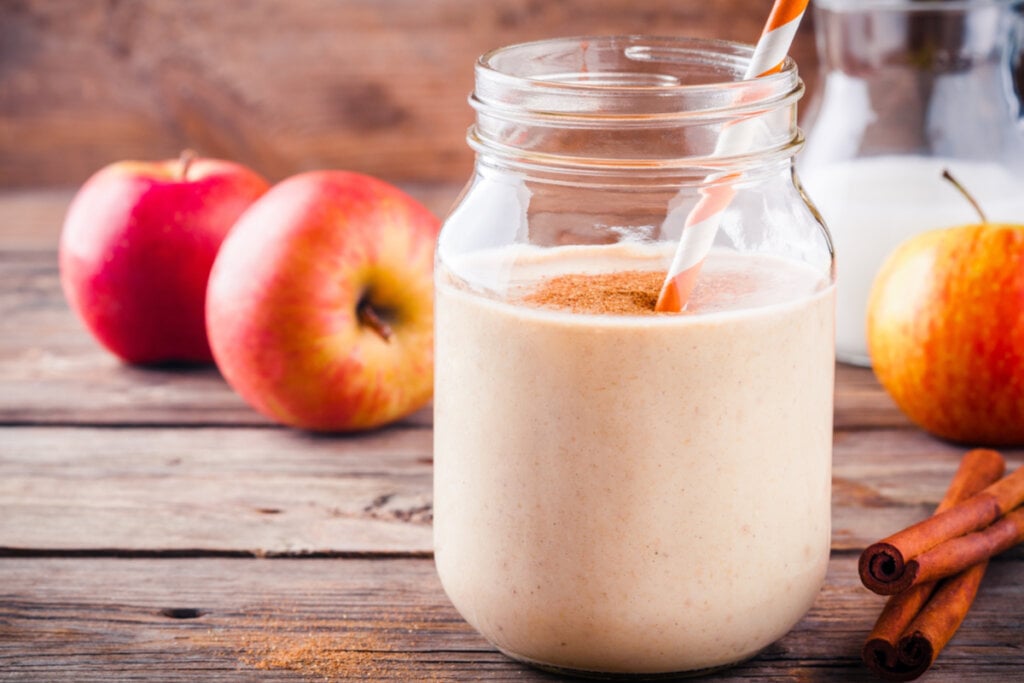 Copo com vitamina de maçã e canela em pó por cima. Ao fundo, há maçã e, ao lado do copo, canela em pau
