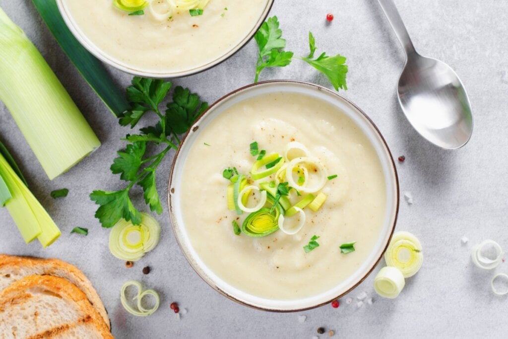 Recipiente branco com sopa de alho-poró em cima de uma mesa cinza com pedaços de alho-poró
