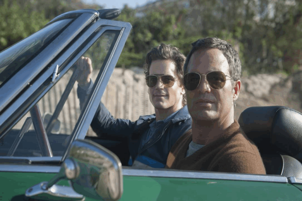 Imagem de dois homens dentro de carro com teto aberto, um deles está usando óculos escuro e uma blusa marrom com camiseta branca e o outro está usando óculos escuro e uma blusa azul.