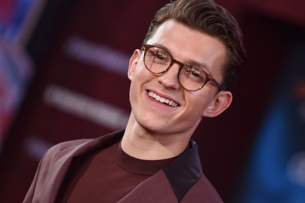 Tom Holland sorrindo, usando óculo de grau e terno cor de vinho