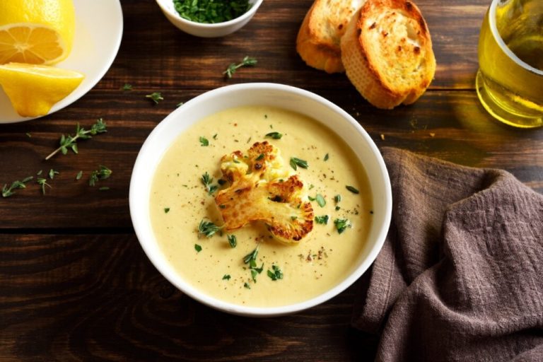 Recipiente branco com caldo de couve-flor decorado com couve-flor tostada e folhas de coentro 