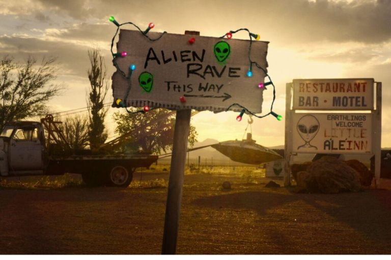 Capa do documentário Desastre Total: Storm Area 51 com uma placa de papelão com pisca-pisca e pedras e caminhões ao fundo 