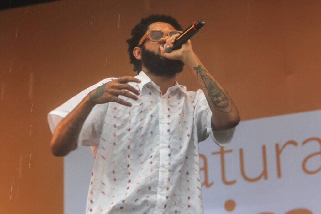 Homem com o cabelo cacheado curto, usando óculos de sol marrom, usando camiseta branca com estampas em flores e com microfone na mão e cantando