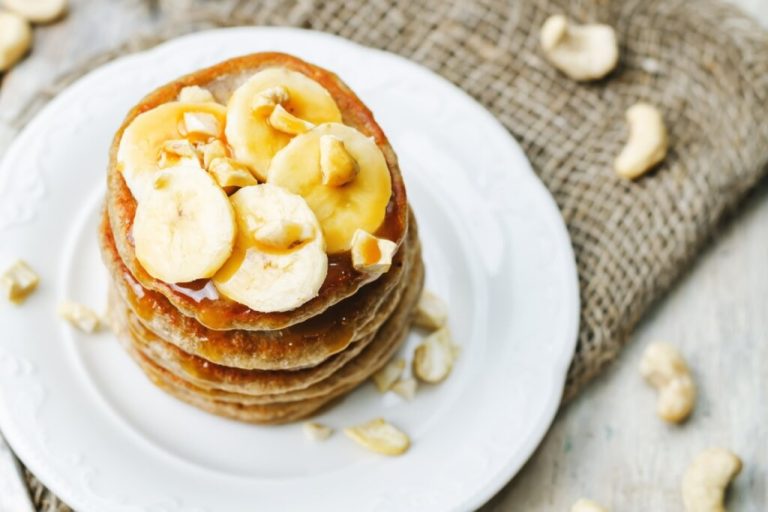 Panquecas de banana empilhadas em um prato branco, regadas com mel e polvilhadas com castanha-de-caju