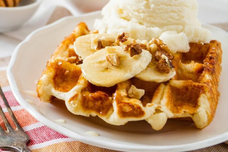 Waffle de banana com nozes em um prato branco com uma bola de sorvete, duas rodelas de banana e nozes em cima 